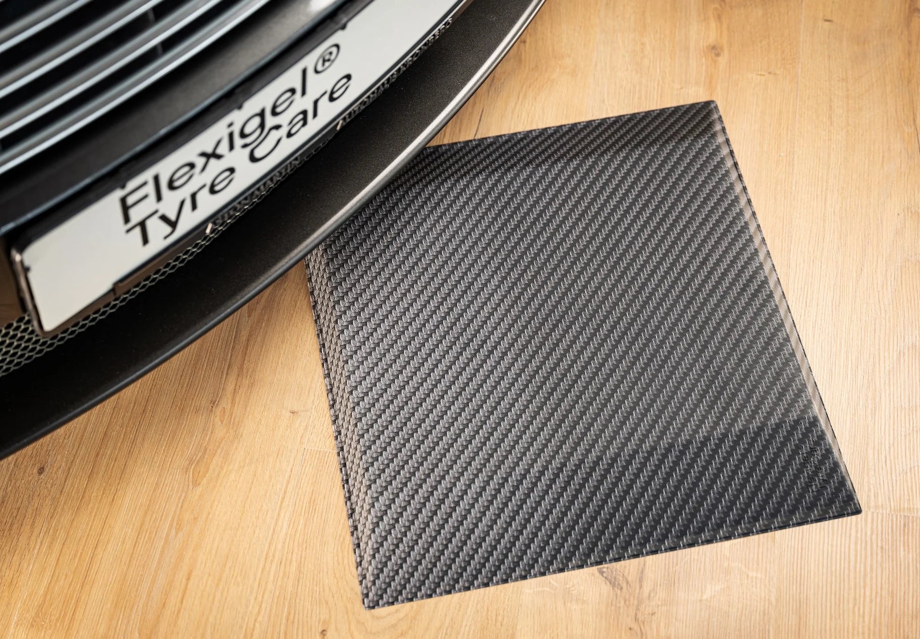 Reifenschoner in Carbon-Optik liegt auf einem Holzboden. Darüber steht ein Fahrzeug mit einem Flexigel® Tyre Care Kennzeichen.